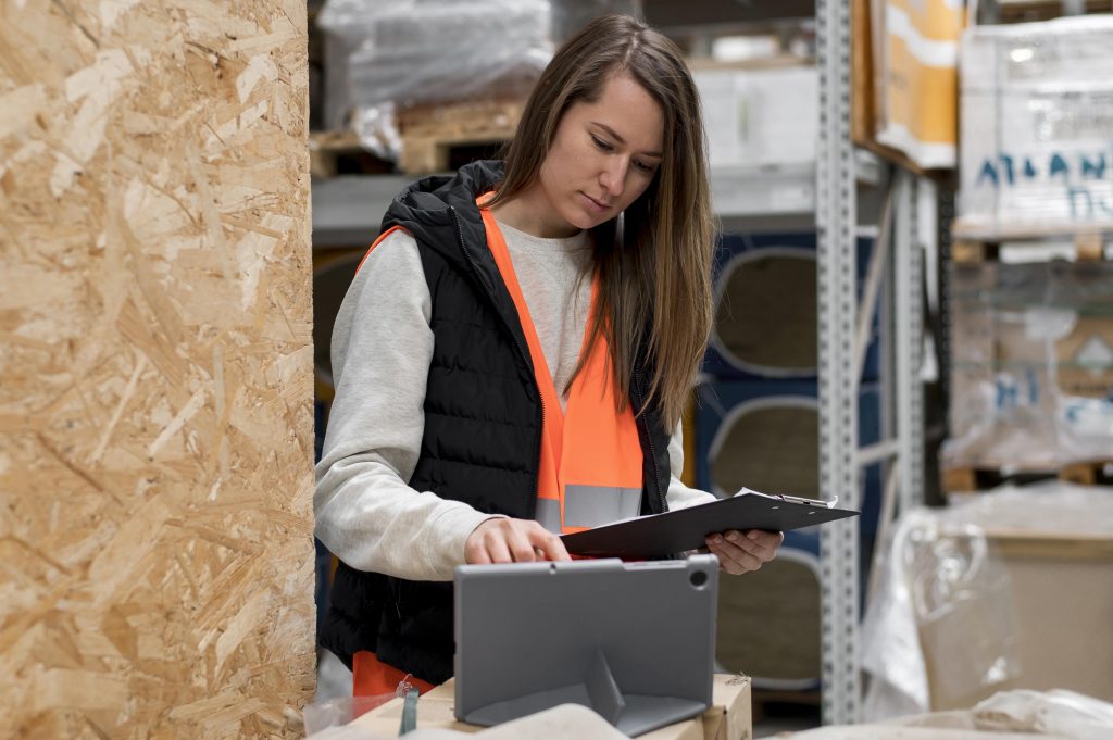 Mulher de colete de segurança laranja em depósito, olhando para prancheta e tablet. Fundo com prateleiras e caixas de madeira.