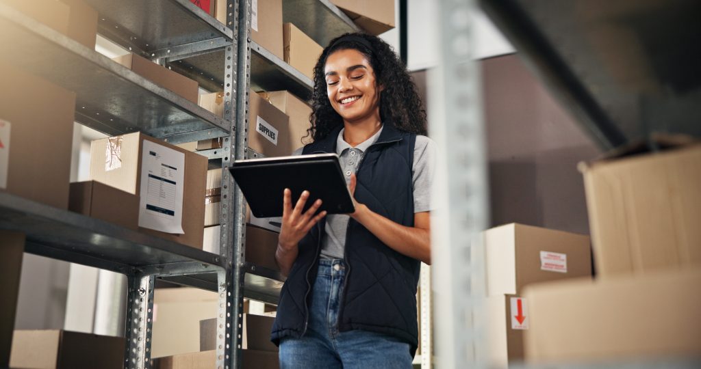Mulher sorridente com colete e tablet no armazém, gerenciando o estoque e pedidos em prateleiras cheias de caixas.