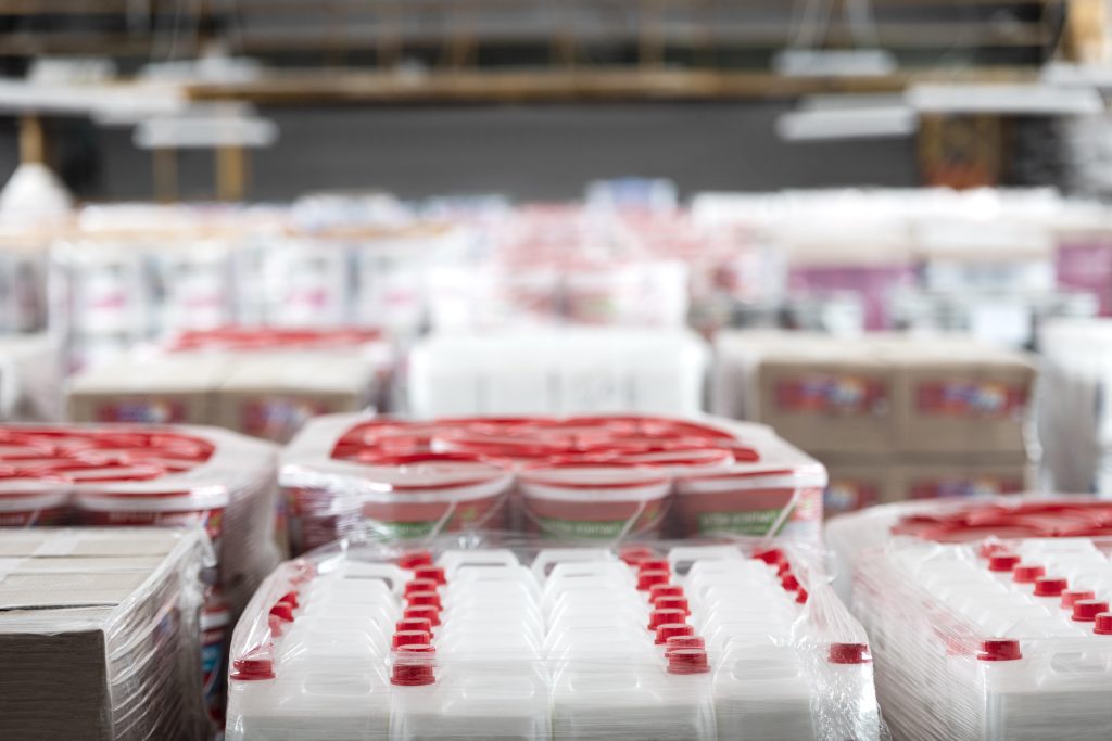 Visão em close-up de produtos embalados em filme plástico no chão de um armazém. Em primeiro plano, galões brancos com tampas vermelhas e, atrás, baldes de plástico empilhados com bordas vermelhas.